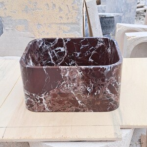 May include: A rectangular, open-top basin made of dark red marble with white veining. The basin has rounded corners and is set on a light-colored wooden surface. The marble has a polished finish.