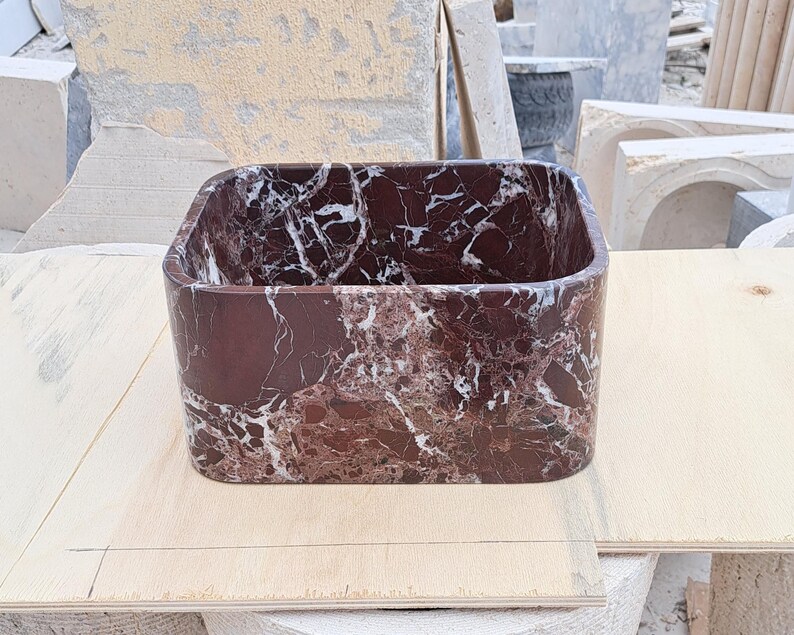 May include: A rectangular, open-top basin made of dark red marble with white veining. The basin has rounded corners and is set on a light-colored wooden surface. The marble has a polished finish.
