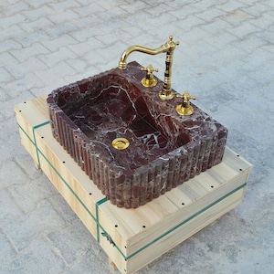 K&ouml;nnte beinhalten: Ein rechteckiges Waschbecken aus rotem Marmor mit einer goldenen Armatur und Armaturen. Das Waschbecken steht auf einer Holzkiste. Das Waschbecken hat eine geriffelte Au&szlig;enseite und einen goldenen Abfluss. Die Armatur ist verziert.