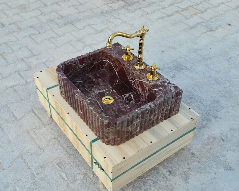 K&ouml;nnte beinhalten: Ein rechteckiges Waschbecken aus rotem Marmor mit einer goldenen Armatur und Armaturen. Das Waschbecken steht auf einer Holzkiste. Das Waschbecken hat eine geriffelte Au&szlig;enseite und einen goldenen Abfluss. Die Armatur ist verziert.