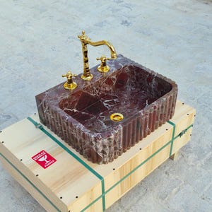 K&ouml;nnte beinhalten: Ein rechteckiges Waschbecken aus rotem Marmor mit goldenen Armaturen und Griffen. Es steht auf einer Holzkiste mit gr&uuml;nen Gurten. Das Innere des Waschbeckens ist tiefrot mit wei&szlig;en Adern.