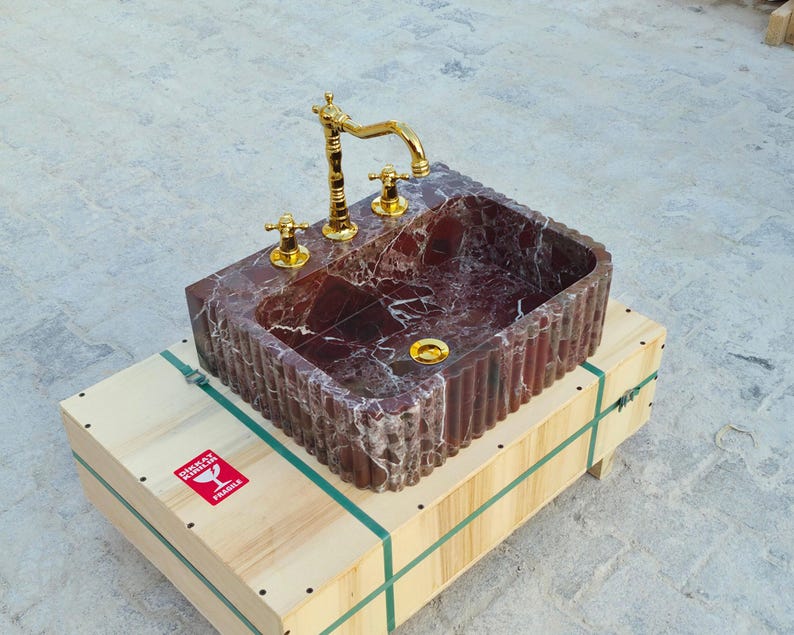 K&ouml;nnte beinhalten: Ein rechteckiges Waschbecken aus rotem Marmor mit goldenen Armaturen und Griffen. Es steht auf einer Holzkiste mit gr&uuml;nen Gurten. Das Innere des Waschbeckens ist tiefrot mit wei&szlig;en Adern.