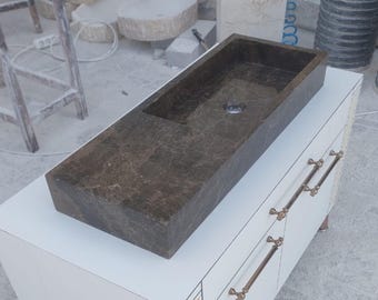 May include: A dark brown marble sink with a rectangular basin, set on a white cabinet with bronze handles. The sink's surface has a textured appearance. The cabinet features gold decorative elements. The sink's dimensions are approximately 80 cm in length.