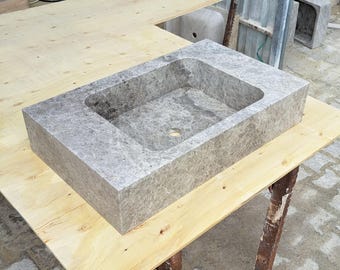 Puede incluir: Un lavabo rectangular de mármol gris con un orificio de desagüe central. El lavabo tiene un interior liso y pulido y un exterior texturizado. El lavabo está sobre una superficie de madera clara, mostrando su apariencia de piedra natural. Es un accesorio de baño.