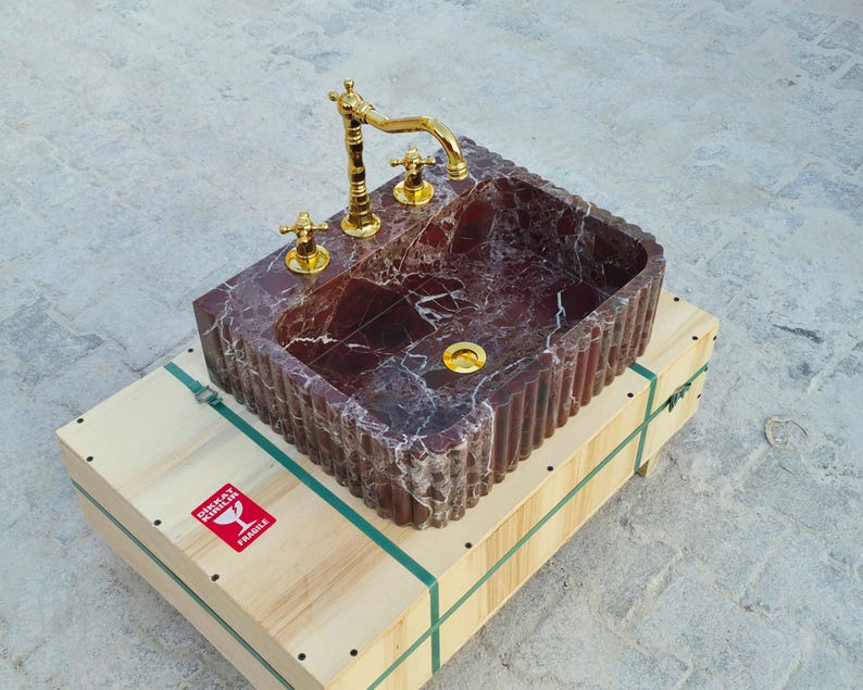 K&ouml;nnte beinhalten: Ein rechteckiges Waschbecken aus burgunderfarbenem Marmor mit goldfarbenen Armaturen und einem geriffelten Rand. Es steht auf einer Holzkiste mit gr&uuml;nen Gurten und einem "Fragile" Aufkleber. Die Ma&szlig;e des Waschbeckens sind nicht angegeben.