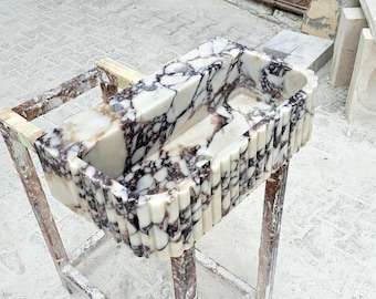 Puede incluir: Un lavabo rectangular de mármol con exterior estriado. El lavabo presenta una base blanca con vetas moradas oscuras y marrones. Está sostenido por un marco de madera. El interior del lavabo es liso, mientras que el exterior tiene un diseño texturizado y acanalado.