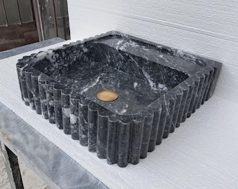 May include: A black marble sink with a fluted edge and a gold drain. The sink is sitting on a white surface.