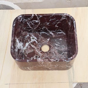 May include: A rectangular, burgundy marble sink with white veining. The sink has a rounded interior and a circular drain hole. The sink is set on a light-colored wooden surface. This bathroom fixture is a stylish addition to any bathroom.