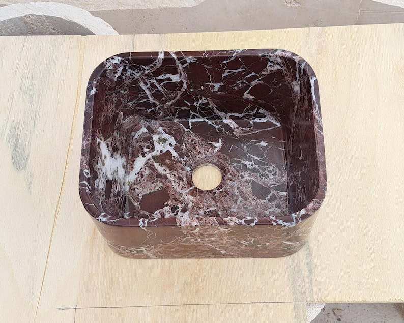 May include: A rectangular, burgundy marble sink with white veining. The sink has a rounded interior and a circular drain hole. The sink is set on a light-colored wooden surface. This bathroom fixture is a stylish addition to any bathroom.