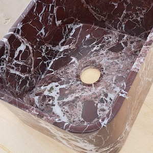 May include: A rectangular, burgundy marble sink with white veining. The sink has a smooth, polished surface and a circular drain opening. The design features a natural stone pattern, suitable for bathroom or kitchen decor.