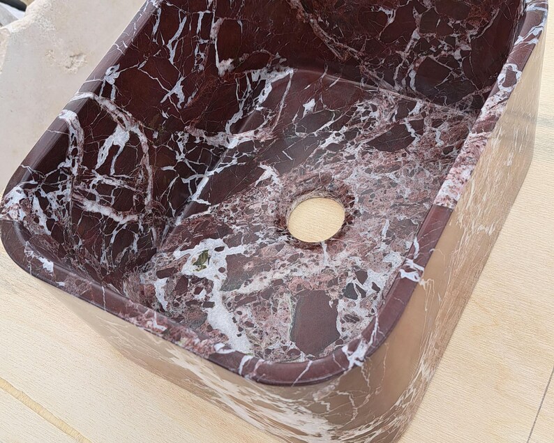 May include: A rectangular, burgundy marble sink with white veining. The sink has a smooth, polished surface and a circular drain opening. The design features a natural stone pattern, suitable for bathroom or kitchen decor.