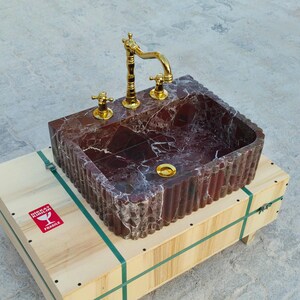 K&ouml;nnte beinhalten: Ein rechteckiges Waschbecken aus rotem Marmor mit goldfarbenen Armaturen. Das Waschbecken steht auf einer Holzkiste. Das Waschbecken hat eine geriffelte Au&szlig;enseite und goldfarbene Wasserh&auml;hne.