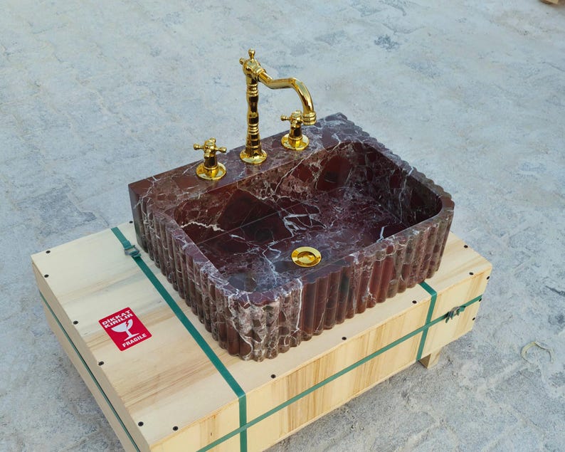 K&ouml;nnte beinhalten: Ein rechteckiges Waschbecken aus rotem Marmor mit goldfarbenen Armaturen. Das Waschbecken steht auf einer Holzkiste. Das Waschbecken hat eine geriffelte Au&szlig;enseite und goldfarbene Wasserh&auml;hne.