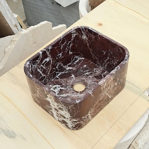 May include: Rectangular bathroom sink made of dark red marble with white veining. The sink has rounded corners and a single drain hole. The sink is sitting on a light-colored wooden surface.
