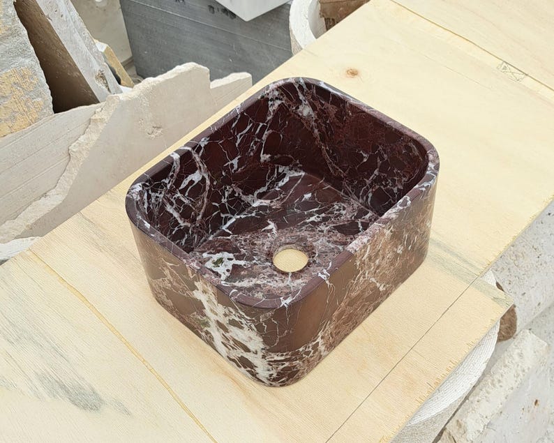 May include: Rectangular bathroom sink made of dark red marble with white veining. The sink has rounded corners and a single drain hole. The sink is sitting on a light-colored wooden surface.