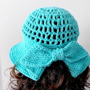 May include: A bright turquoise crochet hat featuring an open-weave pattern on the crown and a solid, tightly stitched brim. A large, prominent crochet bow is attached to the back of the hat, adding texture and detail to the overall design.