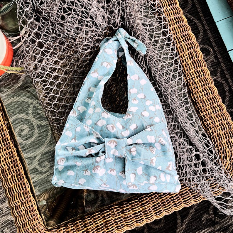 Puede incluir: Un bolso tote azul claro con un estampado floral de flores blancas y grises. El bolso tiene un gran lazo en la parte delantera y un asa anudada. El bolso est&aacute; sobre una superficie tejida.