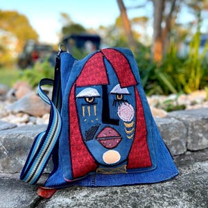 May include: A denim backpack with an abstract face design. The face features red hair, blue skin, and embroidered details in gold, pink, and white. The bag has a striped strap and a red accent at the bottom.