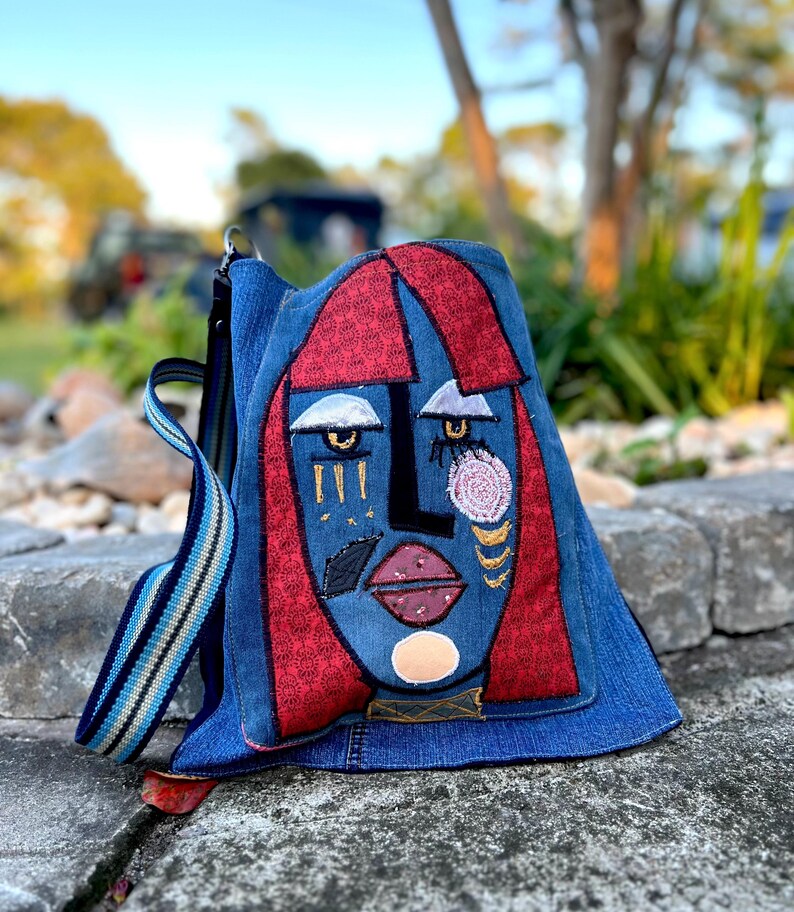 May include: A denim backpack with an abstract face design. The face features red hair, blue skin, and embroidered details in gold, pink, and white. The bag has a striped strap and a red accent at the bottom.