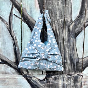 Puede incluir: Bolso tote de tela azul claro con un estampado floral blanco y gris. El bolso tiene un gran lazo y un asa. El bolso est&aacute; colgado en una superficie de madera.