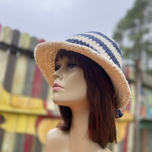 May include: A cream-colored crochet sun hat with a wide brim. The hat features horizontal stripes in a light blue color on the crown. The hat is displayed on a mannequin head with brown hair.