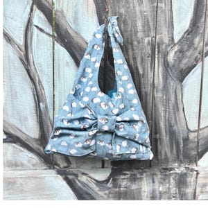 Puede incluir: Un bolso tote azul claro con un estampado floral blanco. El bolso tiene un gran lazo y un asa a juego atada en la parte superior. El bolso cuelga sobre un fondo de madera pintada.