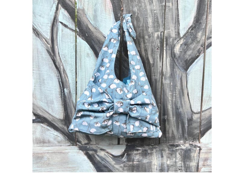 Puede incluir: Un bolso tote azul claro con un estampado floral blanco. El bolso tiene un gran lazo y un asa a juego atada en la parte superior. El bolso cuelga sobre un fondo de madera pintada.