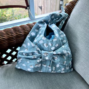 Puede incluir: Un bolso tote de tela azul claro con un estampado floral de flores blancas y tallos marrones. El bolso tiene un gran lazo en la parte delantera y un asa en la parte superior. El bolso est&aacute; sobre un coj&iacute;n azul claro.