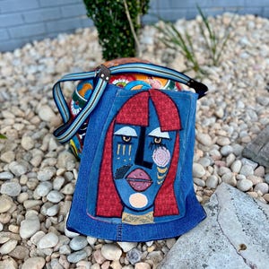 May include: A denim shoulder bag with a colourful patterned body and a flap featuring an abstract face design. The face has red hair, blue skin, and gold accents. The bag has a striped strap.