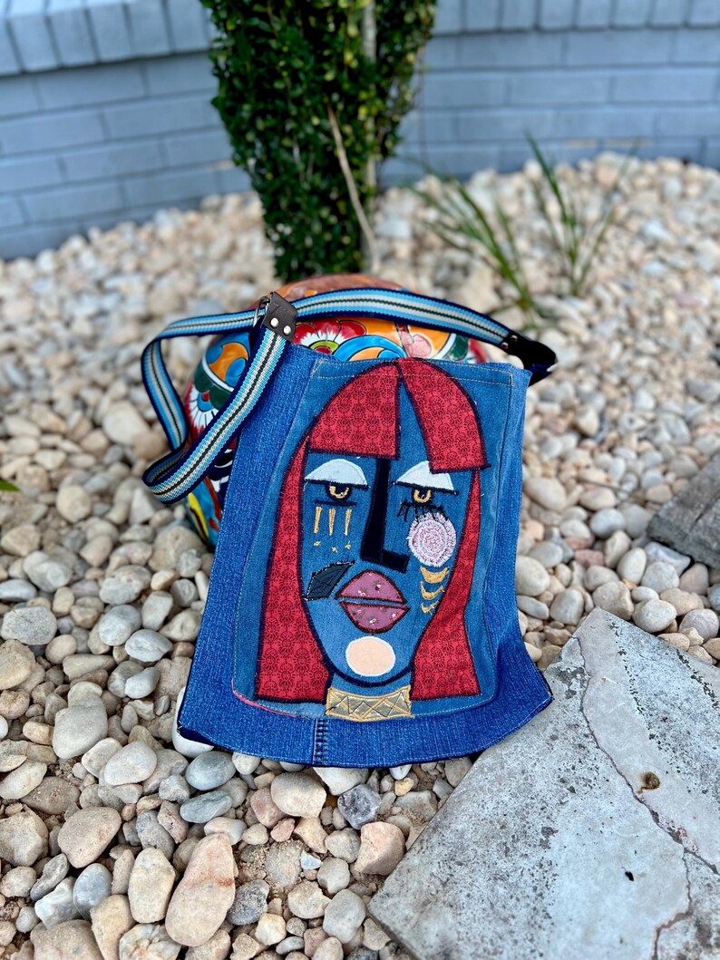 May include: A denim shoulder bag with a colourful patterned body and a flap featuring an abstract face design. The face has red hair, blue skin, and gold accents. The bag has a striped strap.
