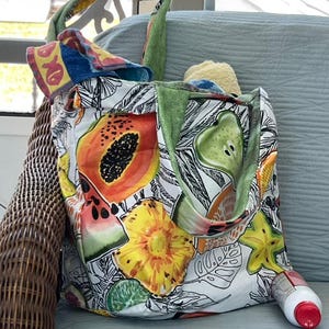 May include: A fabric tote bag featuring a vibrant print of various fruits including papaya, pineapple, watermelon, and starfruit against a black and white botanical background. The bag has green fabric handles and contains folded towels.
