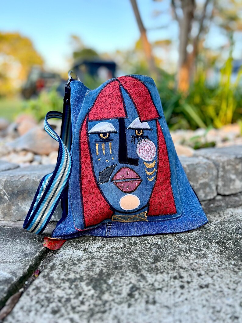 May include: A denim backpack with an abstract face design. The face has red hair, blue skin, and embroidered details like eyes, lips, and gold accents. The backpack features a striped strap and a zip closure. The bag is made of denim.