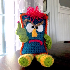Könnte beinhalten: Ein gehäkeltes Plüschtier mit blauem Körper, orangefarbenen und grünen Akzenten und einem mürrischen Gesichtsausdruck. Das Spielzeug hat große weiße Augen, eine braune Nase und einen roten Mund.