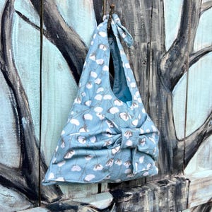 Puede incluir: Bolso tote de tela azul claro con un estampado floral blanco. El bolso tiene un cierre de lazo y una corbata a juego en la parte superior. El bolso est&aacute; colgado en una superficie de madera.