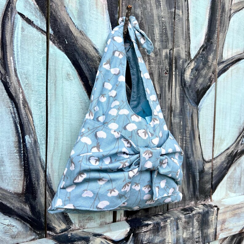 Puede incluir: Bolso tote de tela azul claro con un estampado floral blanco. El bolso tiene un cierre de lazo y una corbata a juego en la parte superior. El bolso est&aacute; colgado en una superficie de madera.