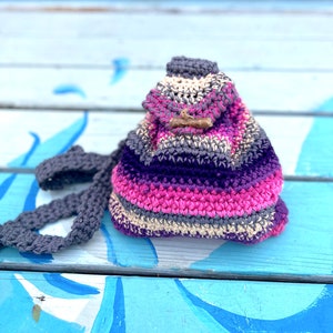 May include: A small, crocheted backpack with a striped pattern in shades of pink, purple, gray, and beige. The backpack has a simple, wooden toggle closure.