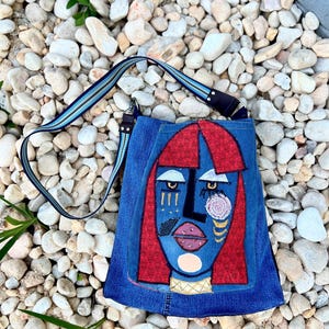 May include: A blue denim shoulder bag with an abstract face design. The face features red hair, a black nose, and pink lips. The bag has a blue and black striped strap and gold embellishments.