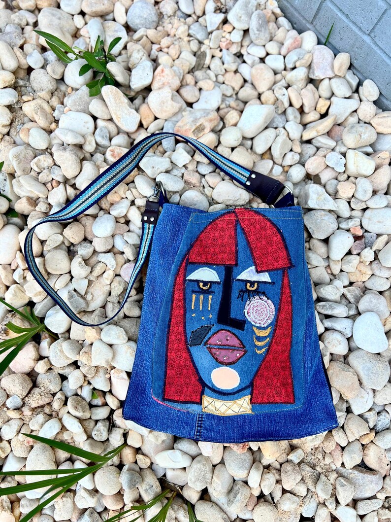May include: A blue denim shoulder bag with an abstract face design. The face features red hair, a black nose, and pink lips. The bag has a blue and black striped strap and gold embellishments.