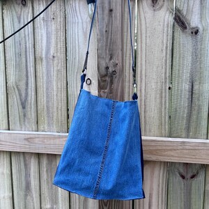 May include: A blue denim crossbody bag with a long strap. The bag has a rectangular shape with a slightly flared bottom. The bag is hanging against a wooden fence.