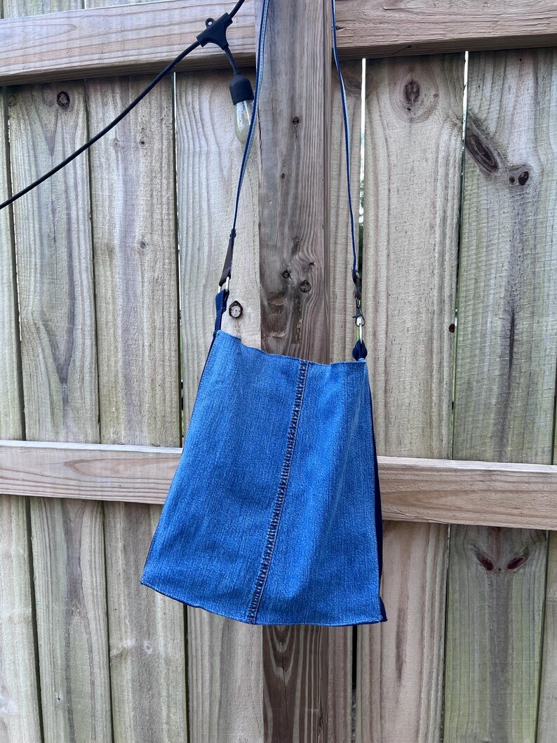May include: A blue denim crossbody bag with a long strap. The bag has a rectangular shape with a slightly flared bottom. The bag is hanging against a wooden fence.