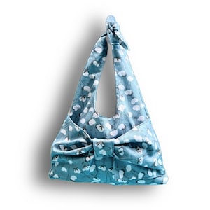 Puede incluir: Bolso tote de color azul claro con un estampado floral blanco. El bolso presenta un gran lazo en la parte delantera y un asa anudada. El bolso de tela est&aacute; dise&ntilde;ado para llevarse al hombro.