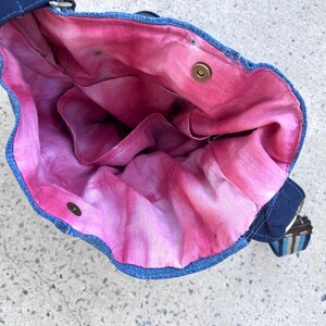 May include: A denim shoulder bag with a vibrant pink and red tie-dye interior. The bag features a dark blue exterior, a matching strap, and a metal snap closure. The interior has multiple pockets.