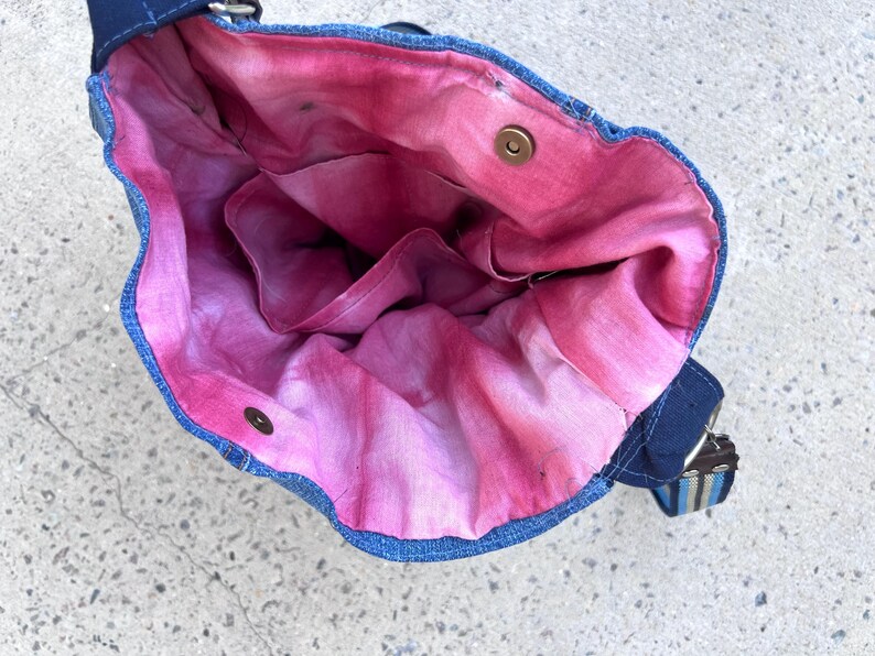 May include: A denim shoulder bag with a vibrant pink and red tie-dye interior. The bag features a dark blue exterior, a matching strap, and a metal snap closure. The interior has multiple pockets.
