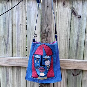 May include: A blue denim crossbody bag with a distinctive face design. The face has red hair, blue skin, and fabric embellishments. The bag has a long, thin strap and is hanging against a wooden fence.