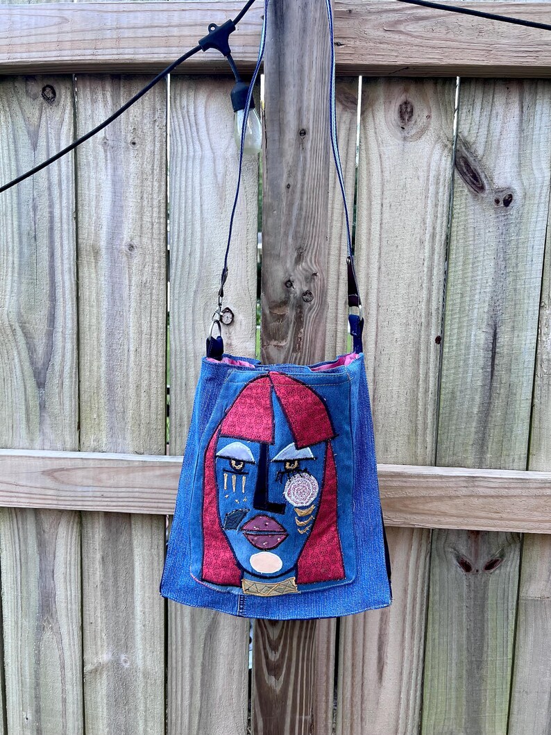 May include: A blue denim crossbody bag with a distinctive face design. The face has red hair, blue skin, and fabric embellishments. The bag has a long, thin strap and is hanging against a wooden fence.