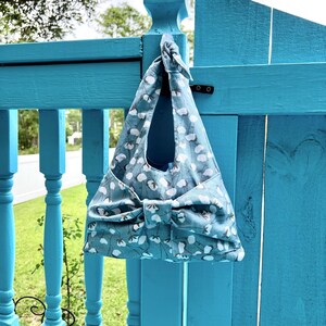 Puede incluir: Un bolso tote azul con un estampado floral blanco cuelga de una valla turquesa. El bolso tiene un lazo grande en la parte delantera y un asa en la parte superior. El bolso est&aacute; hecho de tela y parece hecho a mano.