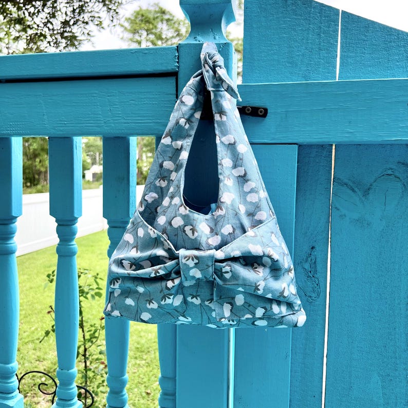 Puede incluir: Un bolso tote azul con un estampado floral blanco cuelga de una valla turquesa. El bolso tiene un lazo grande en la parte delantera y un asa en la parte superior. El bolso est&aacute; hecho de tela y parece hecho a mano.