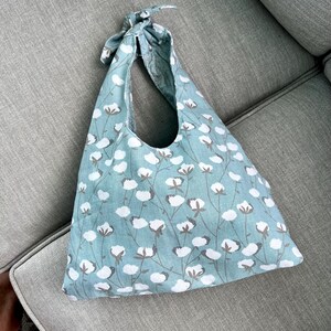 Puede incluir: Bolso tote de color azul claro con un estampado floral blanco y marr&oacute;n. El bolso tiene un asa anudada y est&aacute; hecho de tela. El bolso est&aacute; sobre una superficie gris.