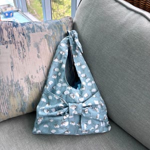 Puede incluir: Bolso tote de color azul claro con un estampado floral blanco y un gran lazo. El bolso es de tela y tiene un asa anudada. Est&aacute; sobre un sof&aacute; gris, junto a un coj&iacute;n estampado.