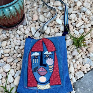 May include: A denim crossbody bag with an abstract face design. The face has red hair, blue skin, and geometric shapes. The bag has a striped strap. The background features pebbles and greenery.